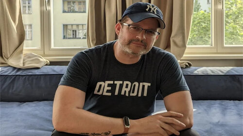 Sascha Pallenberg sitzt in einem Zimmer, trägt ein 'Detroit'-Shirt und eine Brille mit New York Yankees Cap.