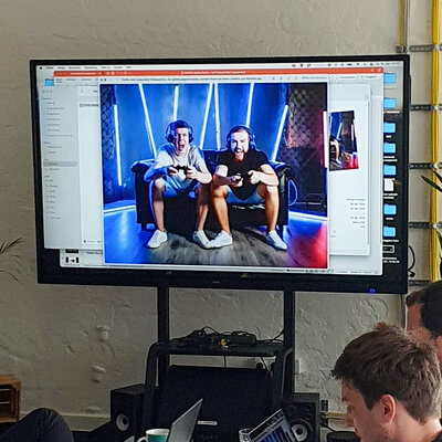 Teilnehmer arbeiten an Laptops, während auf einem Bildschirm zwei Männer beim Gaming gezeigt werden.