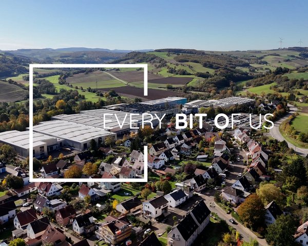 Luftaufnahme einer ländlichen Gegend mit Wohnhäusern im Vordergrund und einer großen Industrieanlage im Hintergrund, überlagert mit dem Schriftzug 'EVERY BIT OF US' in einem weißen, stilisierten Rahmen.