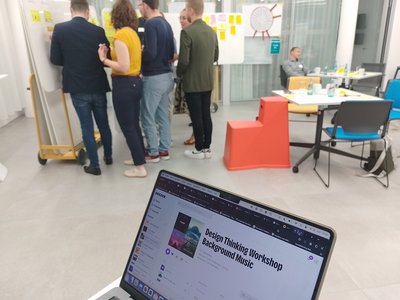 Laptop mit Deezer Screen, Design Thinking Workshop Backround Music wird gespielt, im Hintergrund stehen 6 Personen an einem Whiteboard