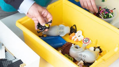 Lego Serious Play Workshop Kiste: Eine Hand nimmt einen Lego-Baustein aus einer gelben Kiste voller verschiedener Teile.