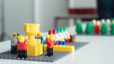 Lego Serious Play Workshop Pokal: Ein aus gelben Lego-Bausteinen gebauter Pokal, den zwei Lego-Figuren in die Höhe halten.